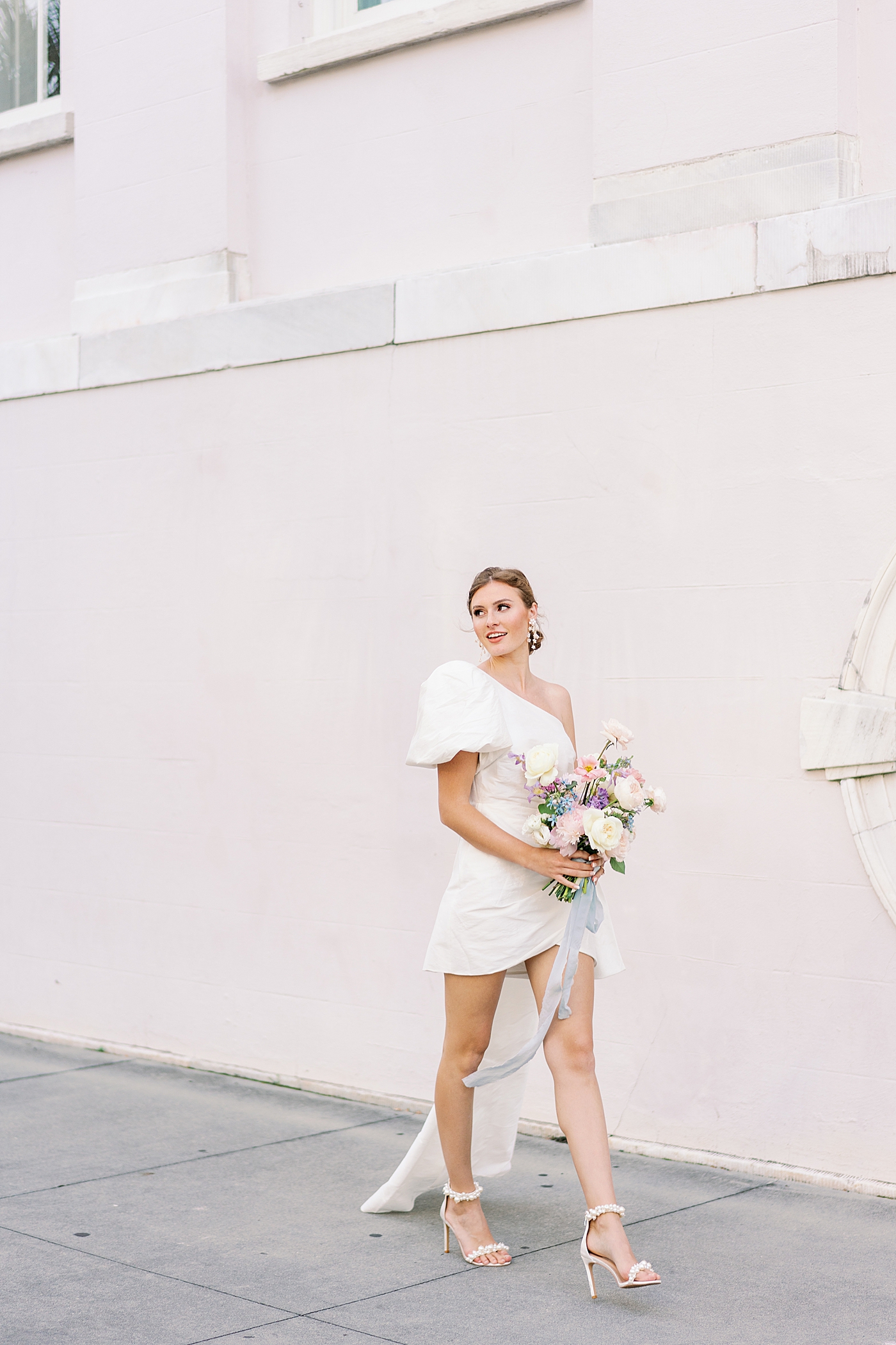 Bride in short gown walking near pink wall holding pastel bouquet | Images by Annie Laura Photo